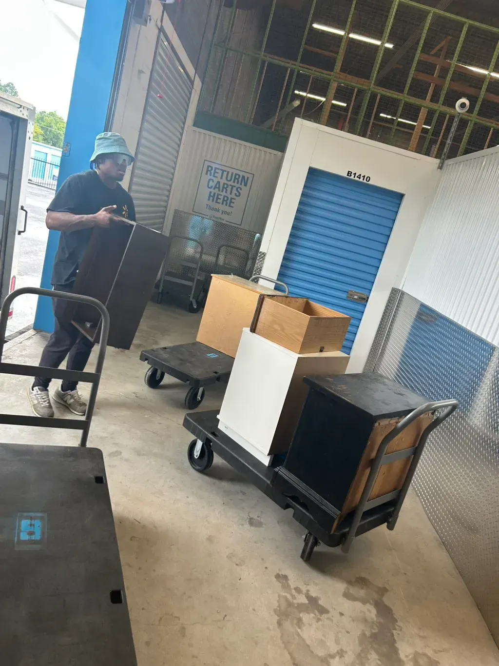A person in a bucket hat carries a dark box toward a storage unit, passing carts loaded with boxes and furniture.