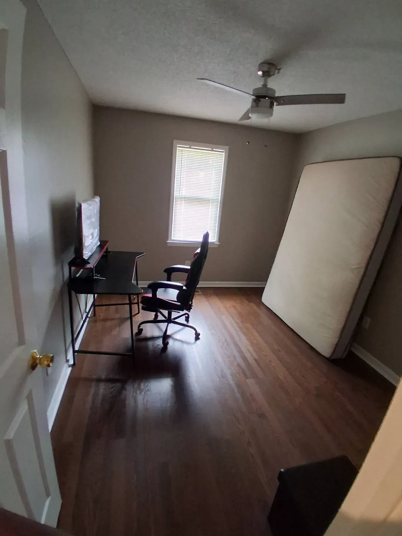 A room with dark wood flooring, a desk with a monitor and chair by a window, and a large mattress leaning against a wall.