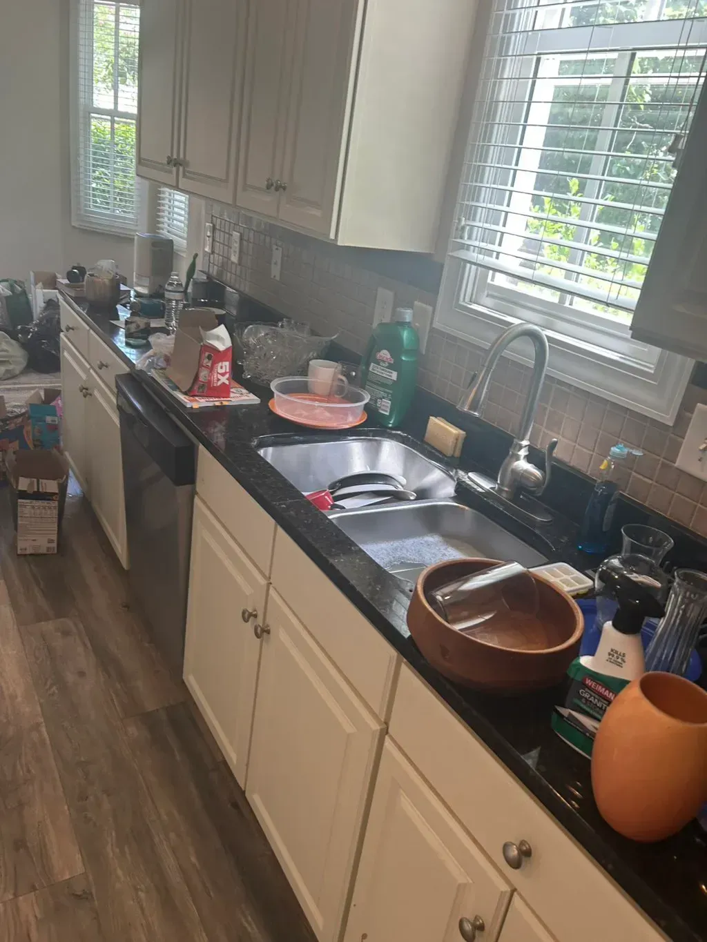A kitchen sink and dark countertop cluttered with dirty dishes, cleaning supplies, and a wooden bowl in a sunny room.