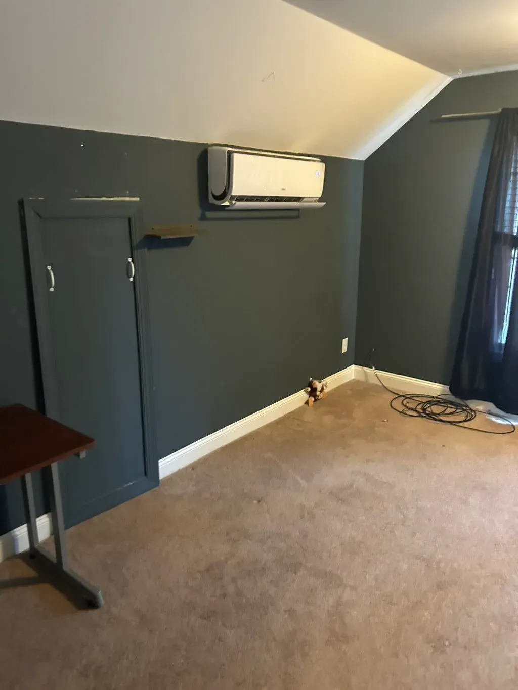 A corner of a room with dark gray walls, beige carpeting, a white mini-split AC unit, and a tall, narrow storage cabinet.