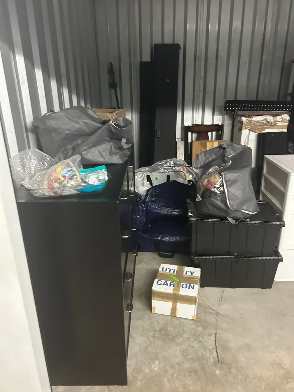 A storage unit containing dark furniture, plastic storage bins, several trash bags, and a cardboard box.