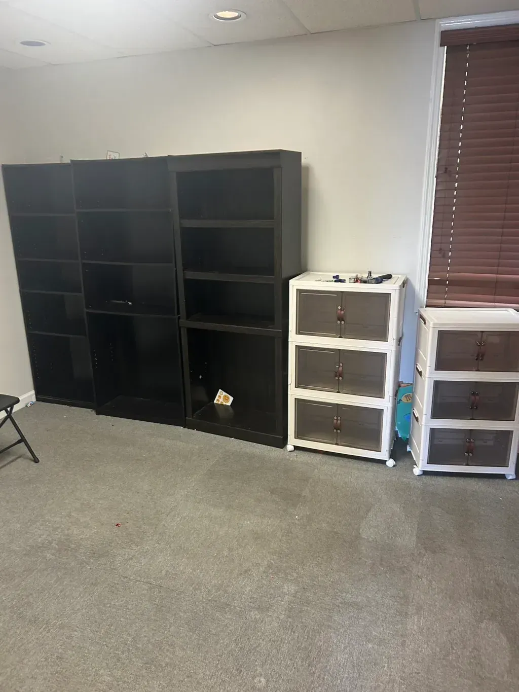 Three black wooden bookshelves stand next to two white plastic drawer units in a room with grey carpet and wooden blinds.