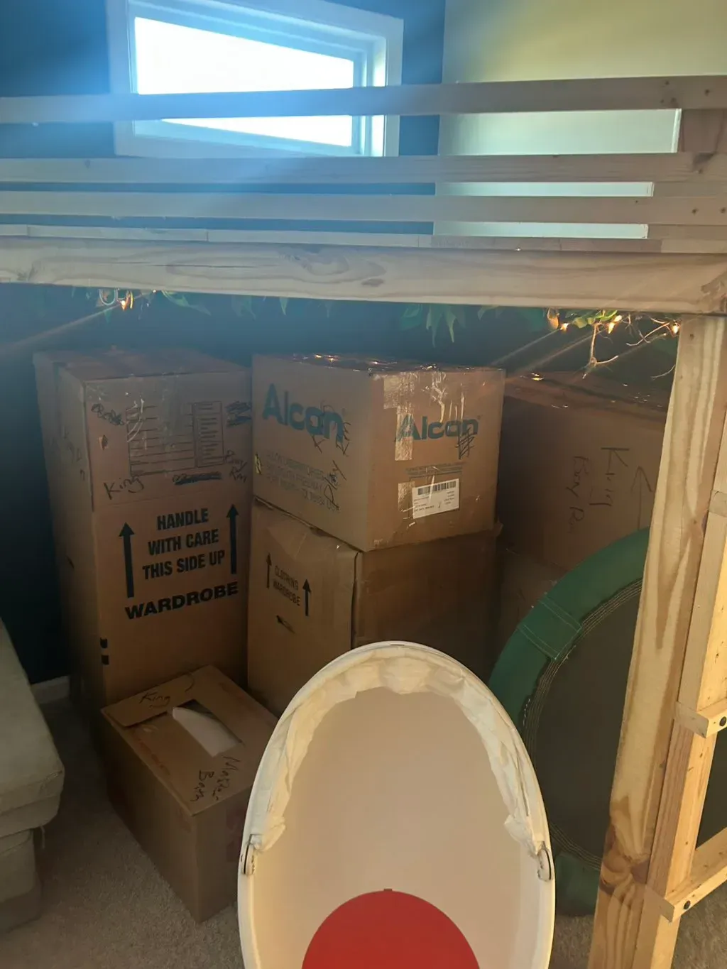 A wooden bunk bed frame stands over cardboard moving boxes and a white, egg-shaped chair with a red seat inside a room.