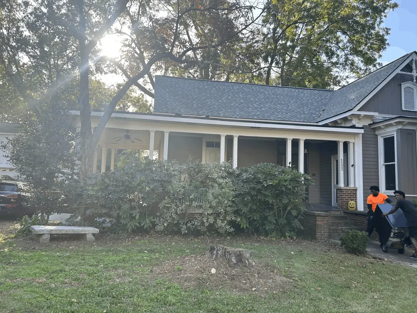 Two people carry a large object onto the front porch of a gray, one-story house with a wide, shaded porch.