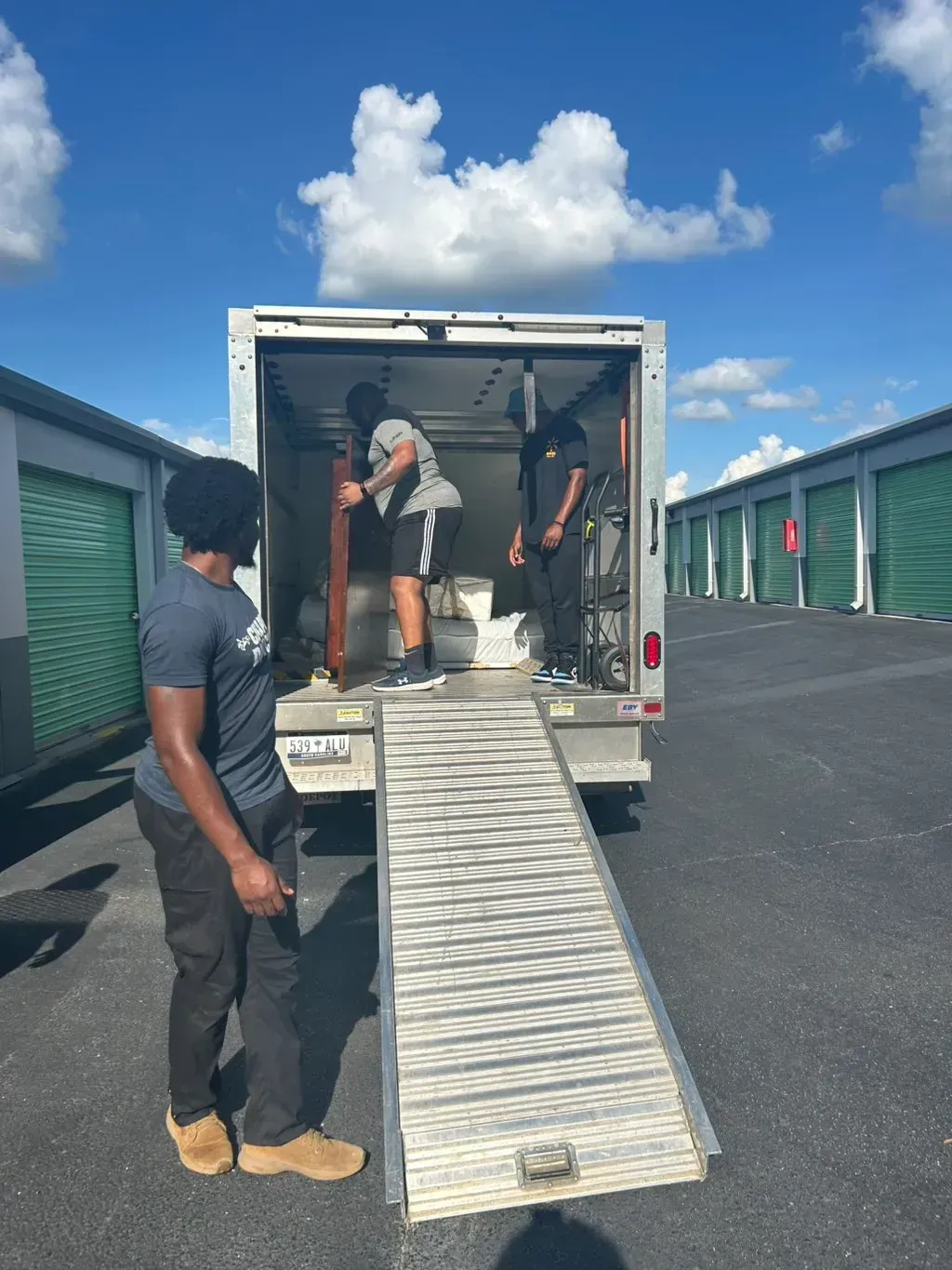 Two people load furniture into a moving truck at a self-storage facility under a blue, cloudy sky.