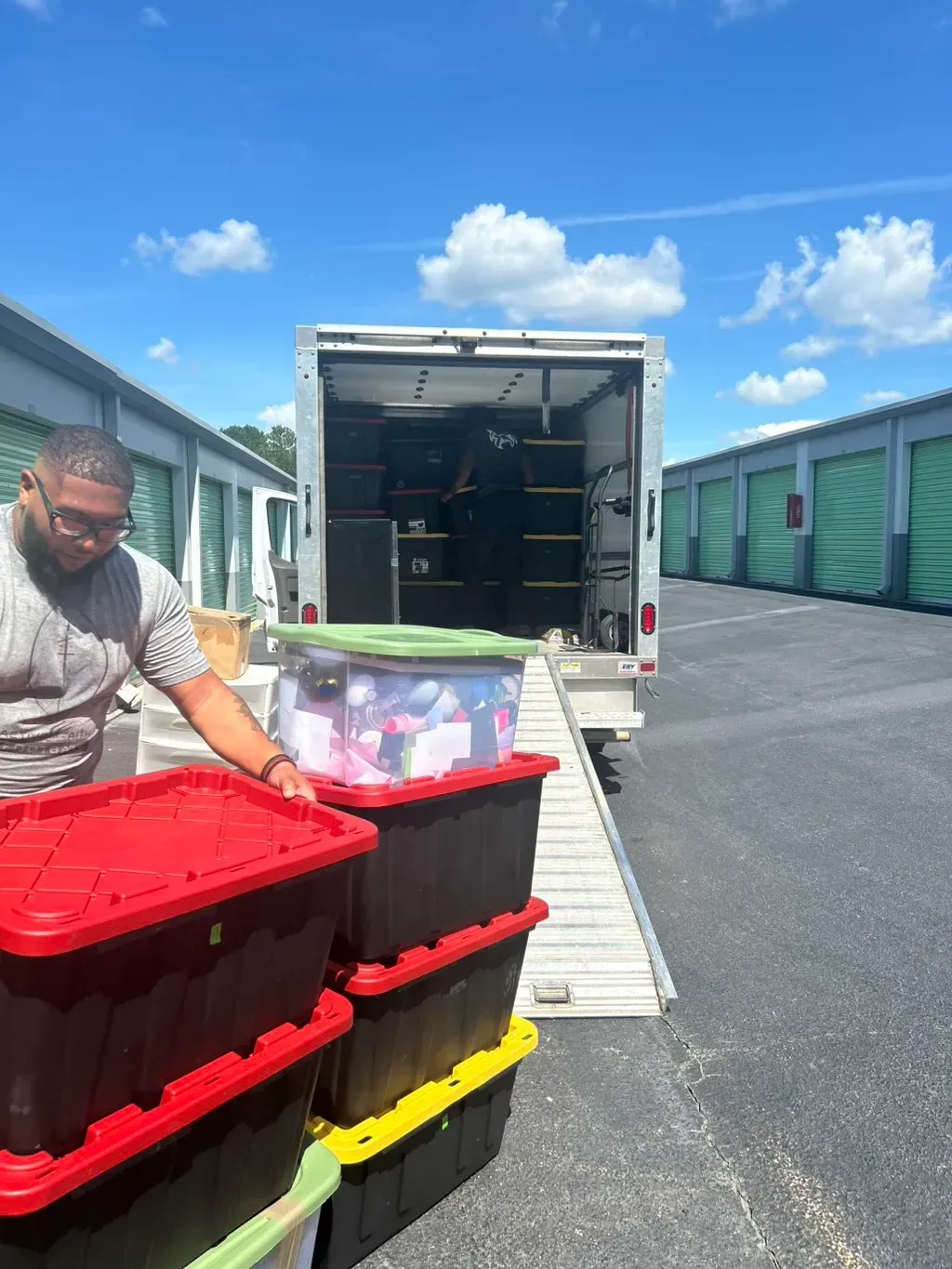 A person loads plastic storage bins into a moving truck at a self-storage facility on a sunny day.