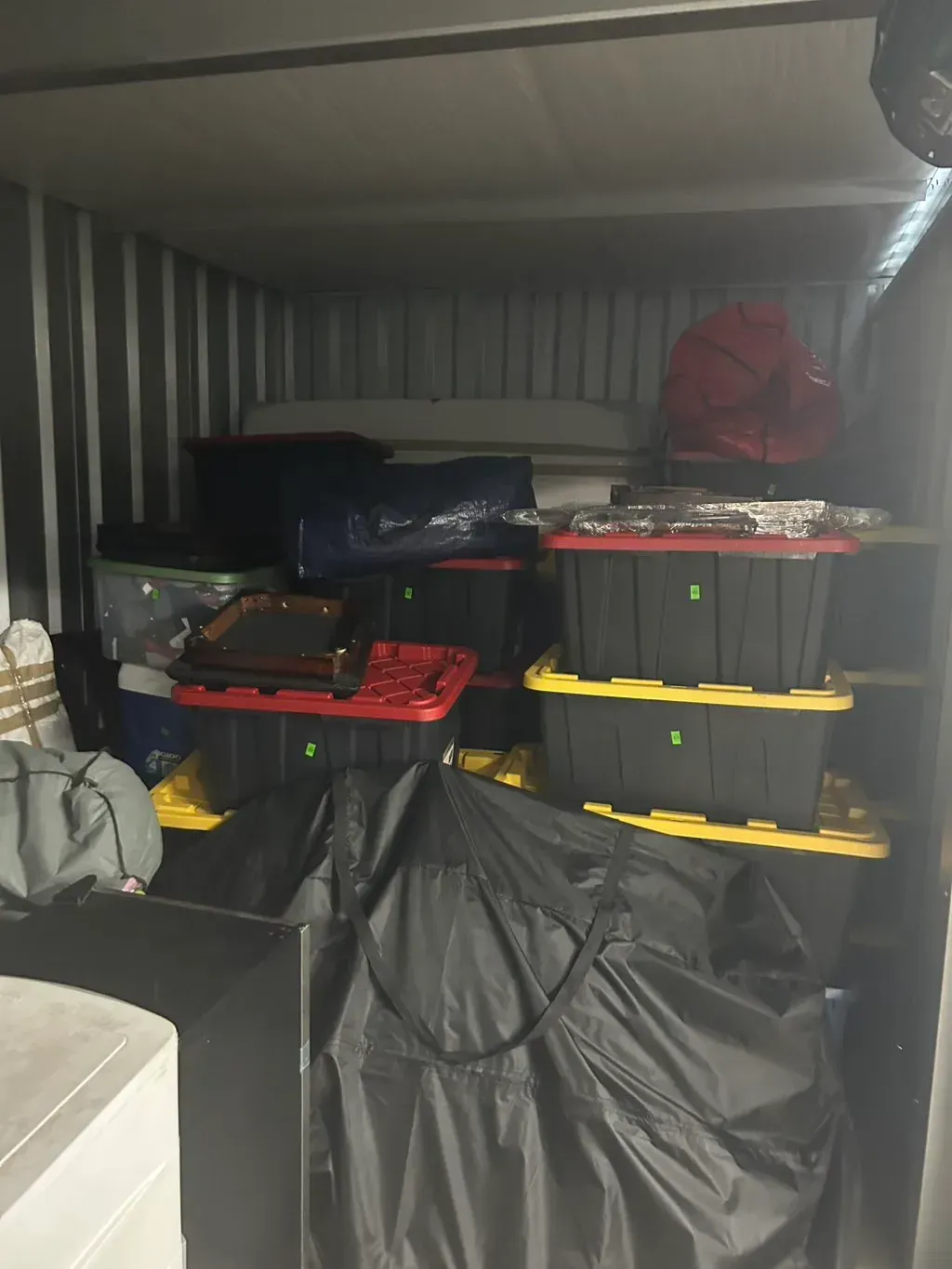 A storage unit filled with stacked black plastic bins with colored lids, a large black duffel bag, and other packed items.
