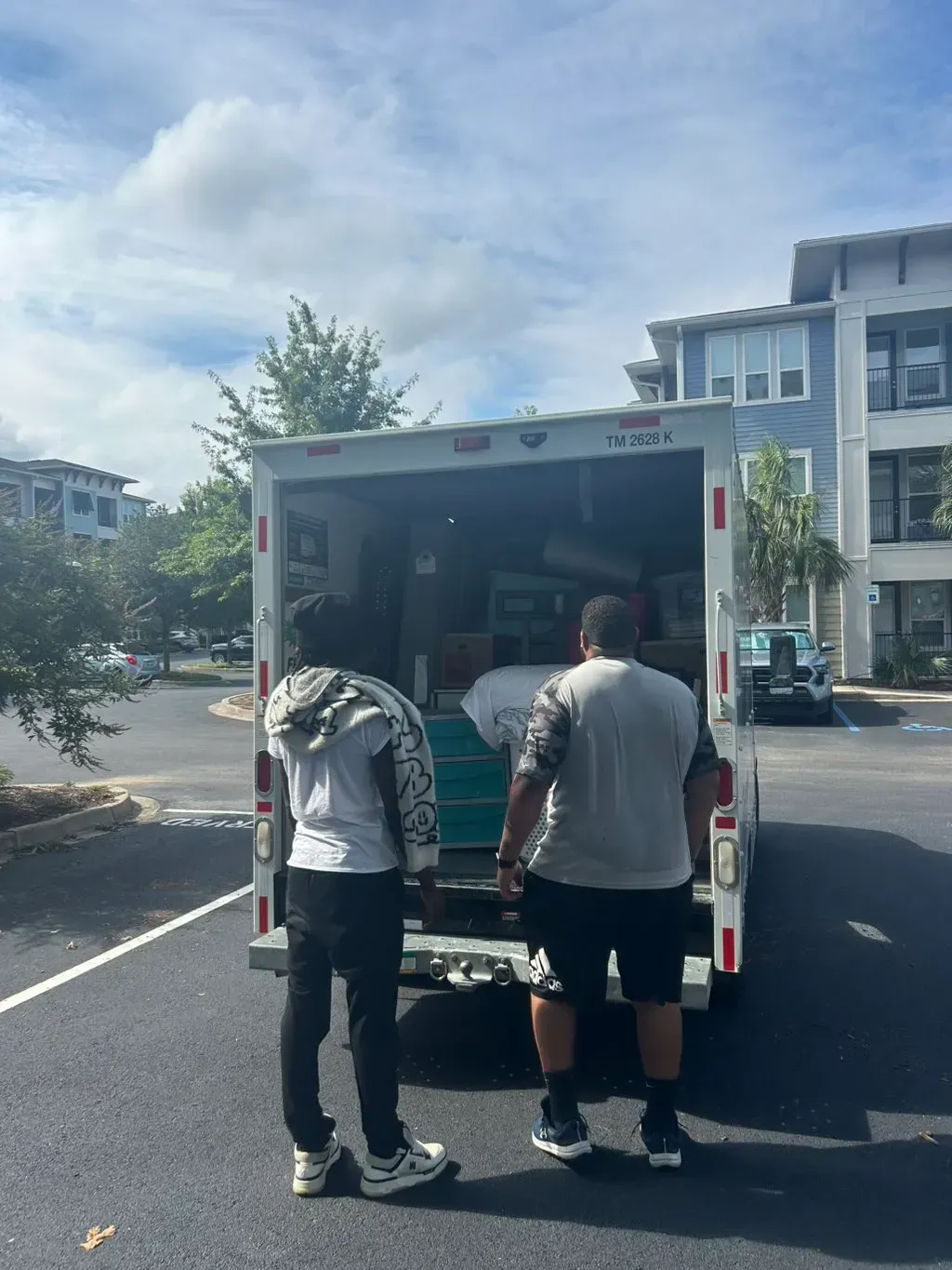 Two people stand at the back of an open moving truck in a parking lot, loading boxes and furniture into the vehicle.