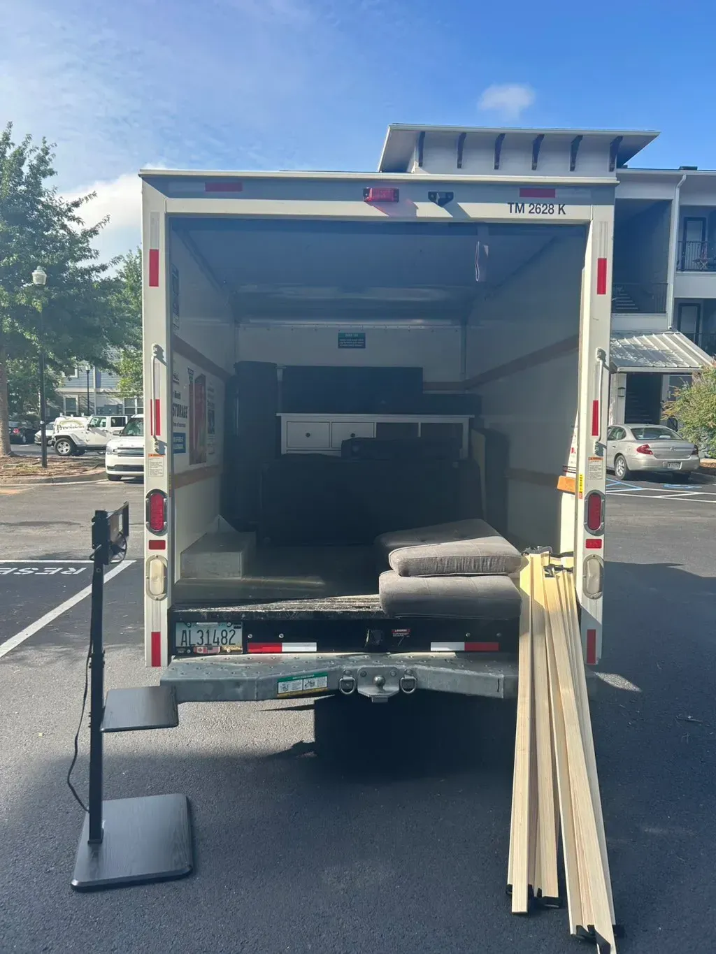 The open rear of a parked moving truck reveals furniture and padding inside, with a floor stand and wooden slats nearby.