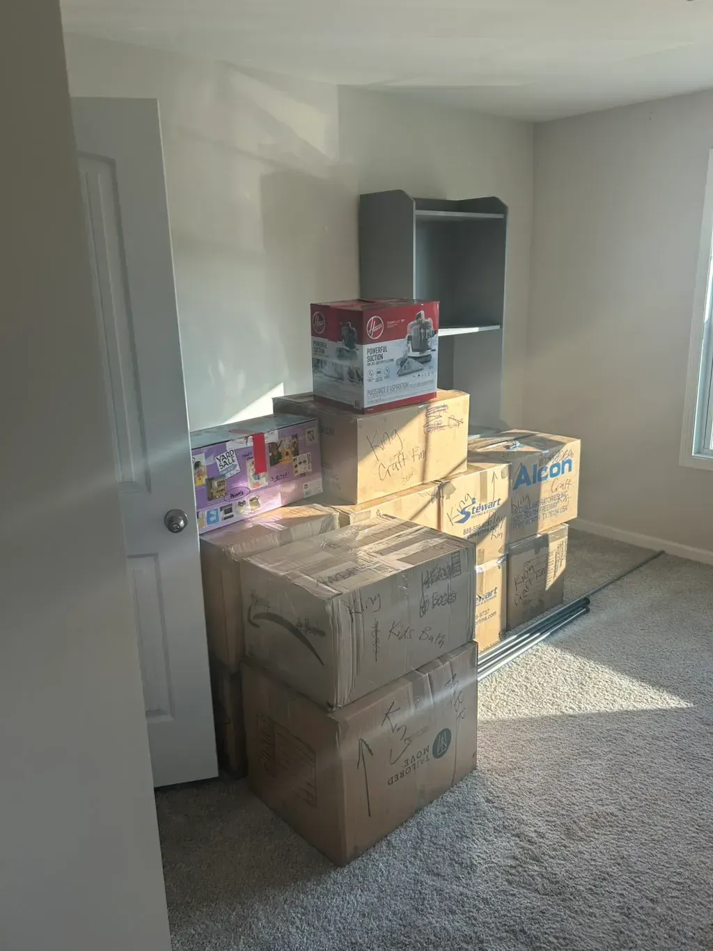 A stack of cardboard moving boxes in a room with a door, a small shelf, and light-colored carpeting.