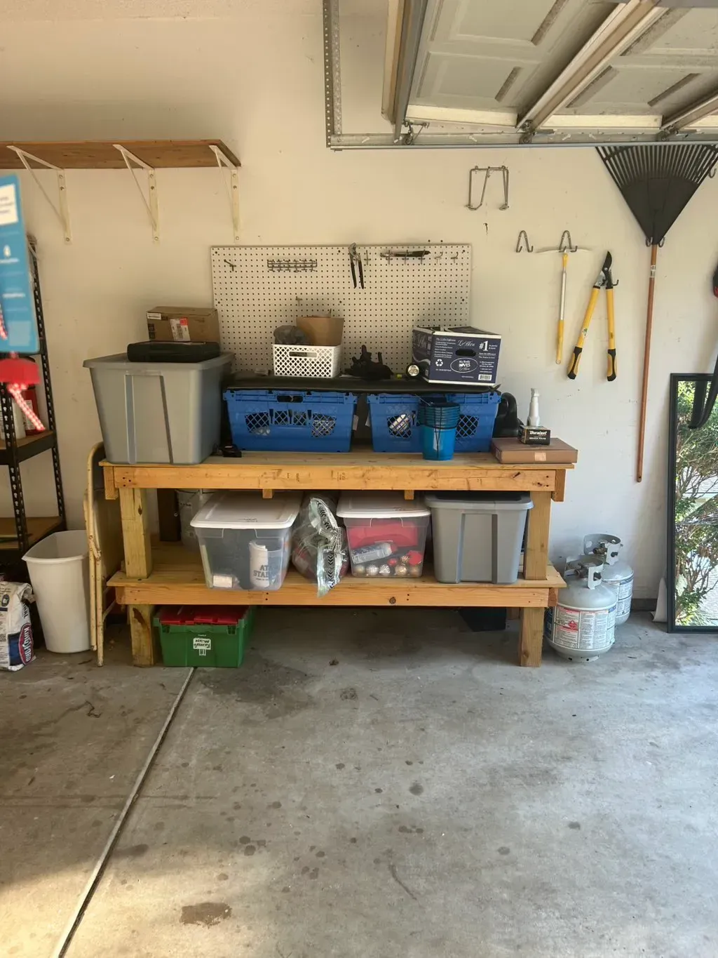 A wooden workbench in a garage filled with plastic storage bins, tools, and supplies, against a white wall with pegboard.