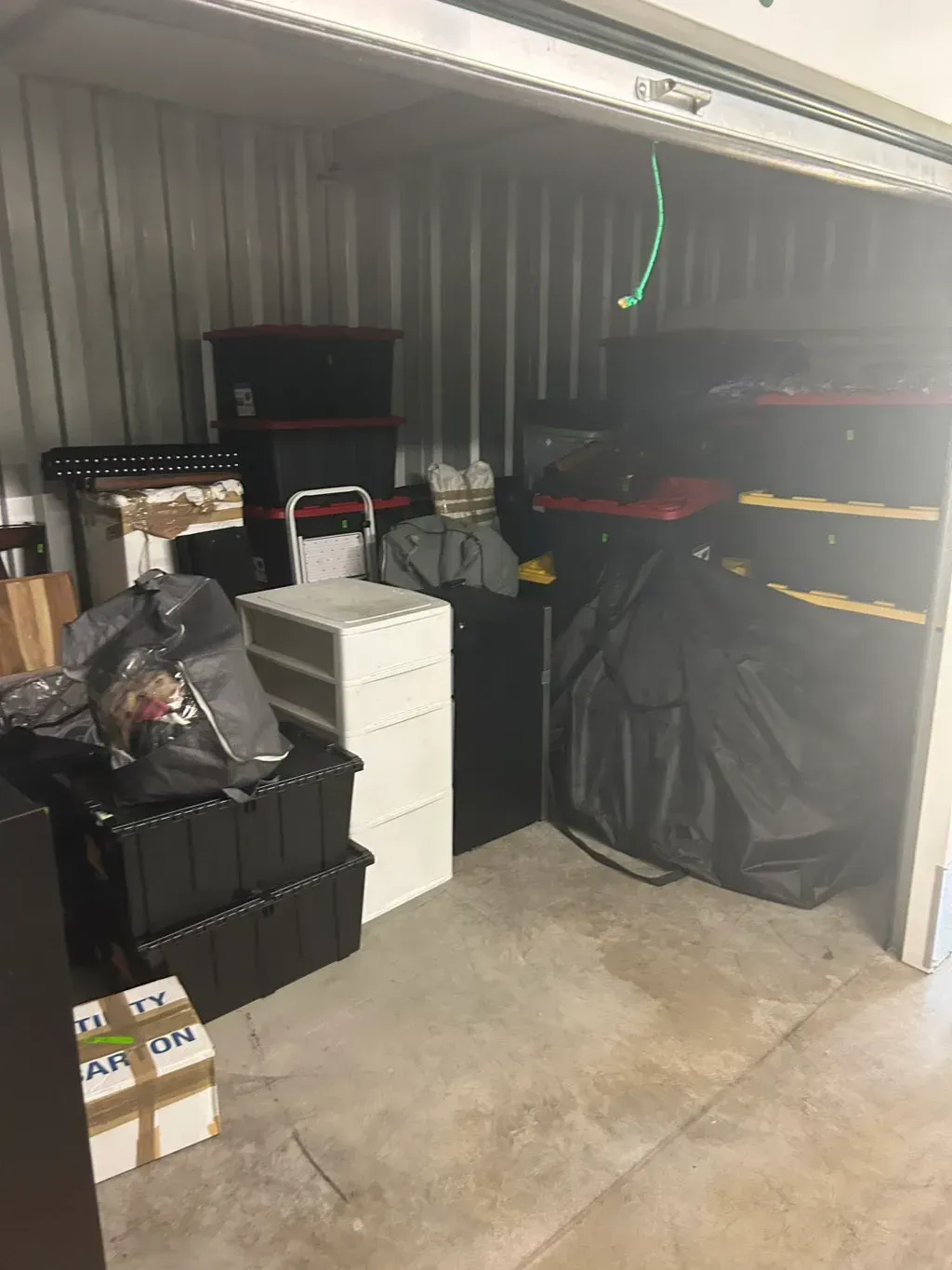 A cluttered storage unit filled with stacked black storage bins, a white plastic drawer unit, and various bags.