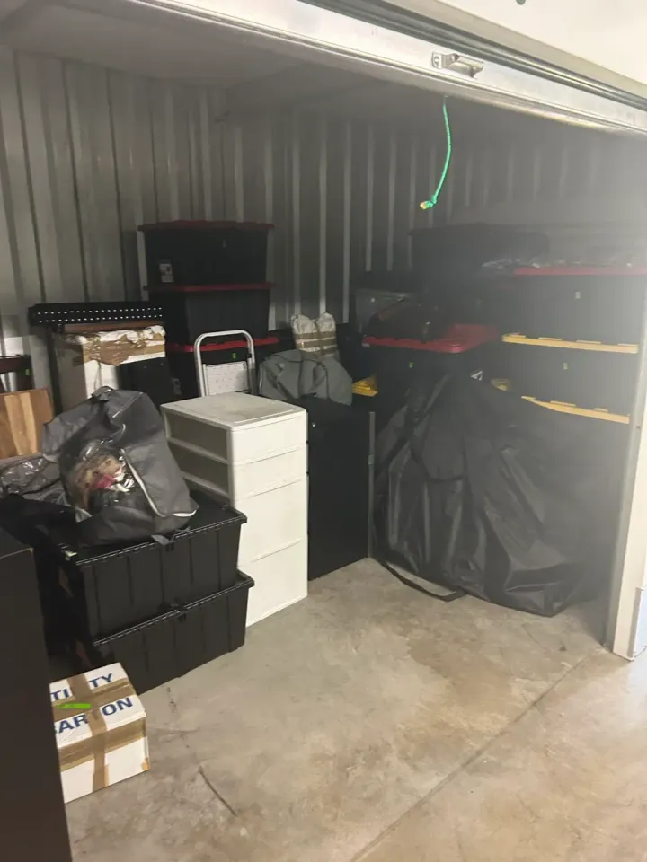 A cluttered storage unit filled with stacked black storage bins, a white plastic drawer unit, and various bags.