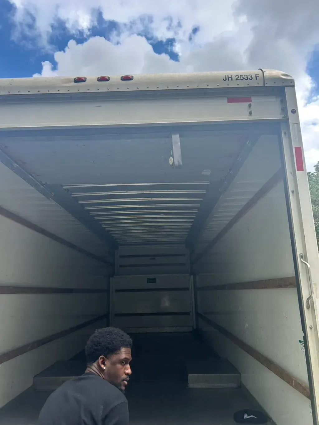 A person stands in front of the open, empty cargo bay of a large moving truck against a blue sky with clouds.