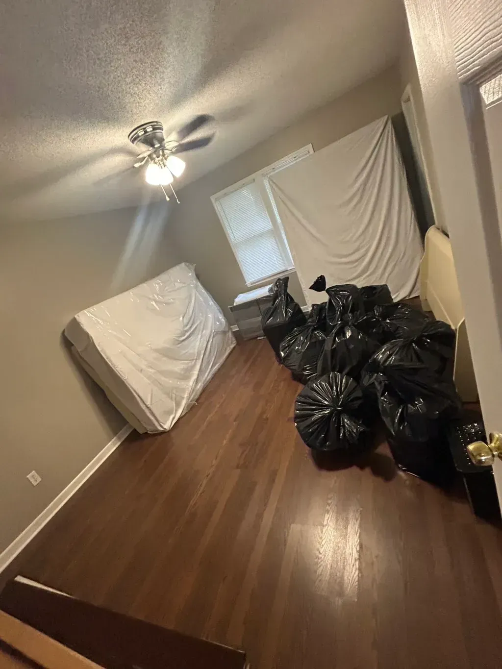 A bedroom with dark wood flooring, a mattress in a protective cover, and a pile of black trash bags on the floor.