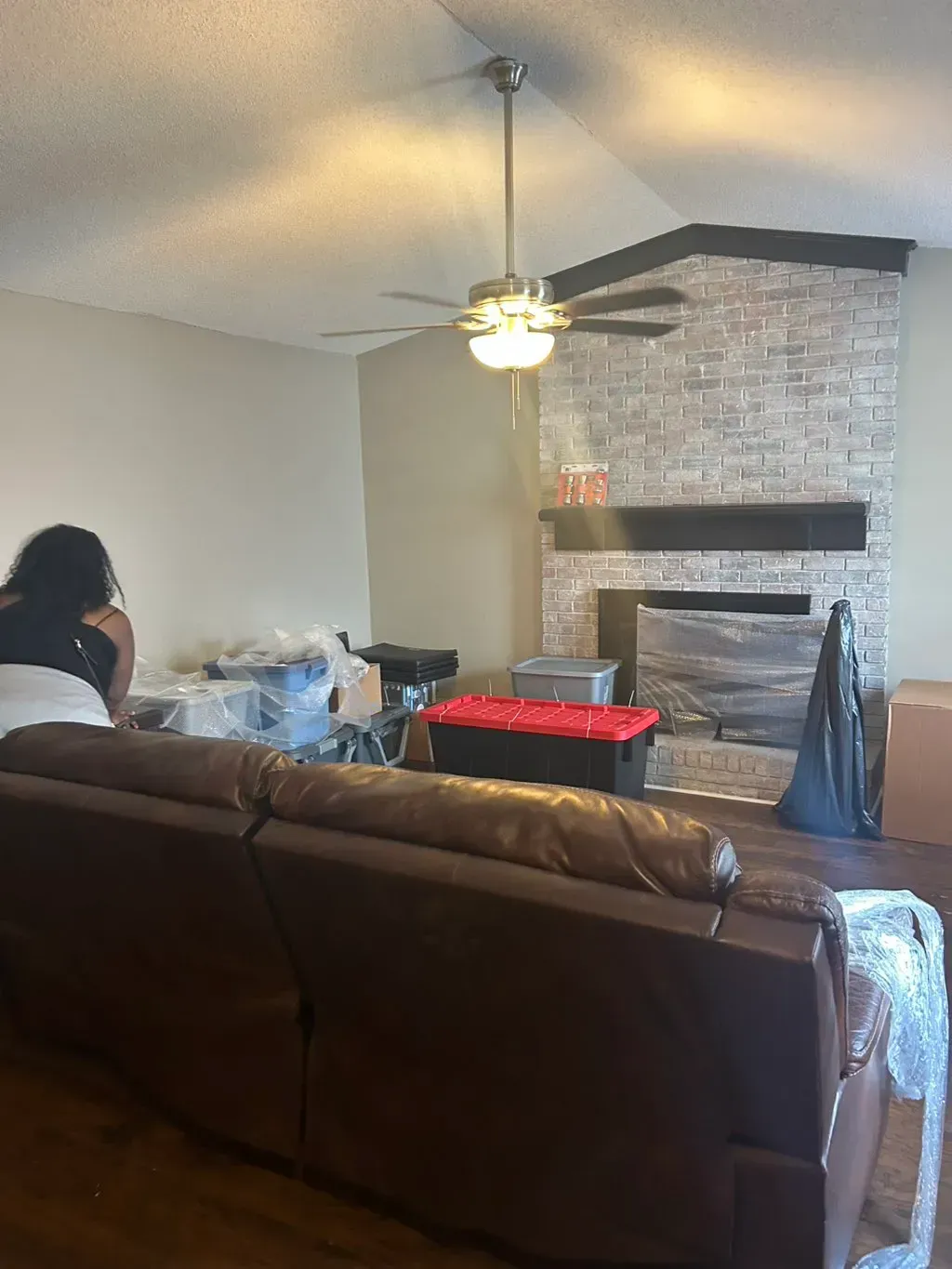 A living room featuring a brown leather sofa, a brick fireplace with a black mantel, and packed moving boxes.