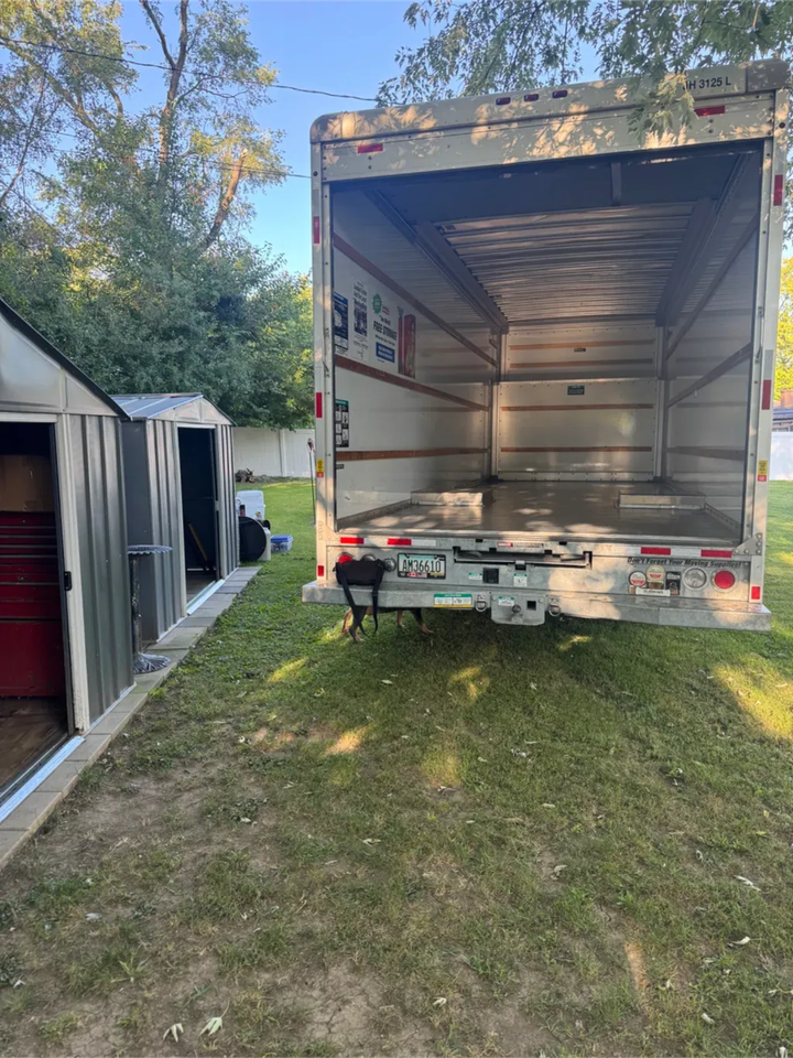 An open cargo trailer parked on grass next to a small, gray metal storage shed on a sunny day.