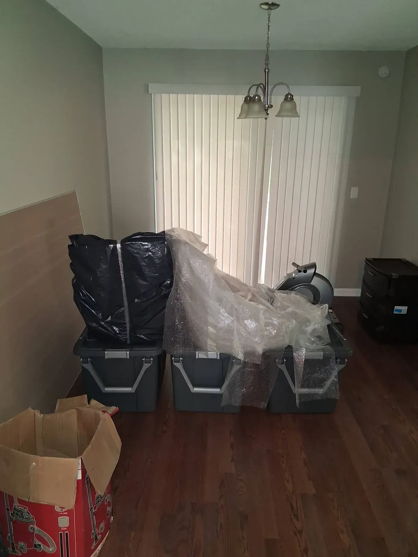 Three gray plastic storage bins filled with wrapped items sit on a wood floor in front of a sliding glass door.