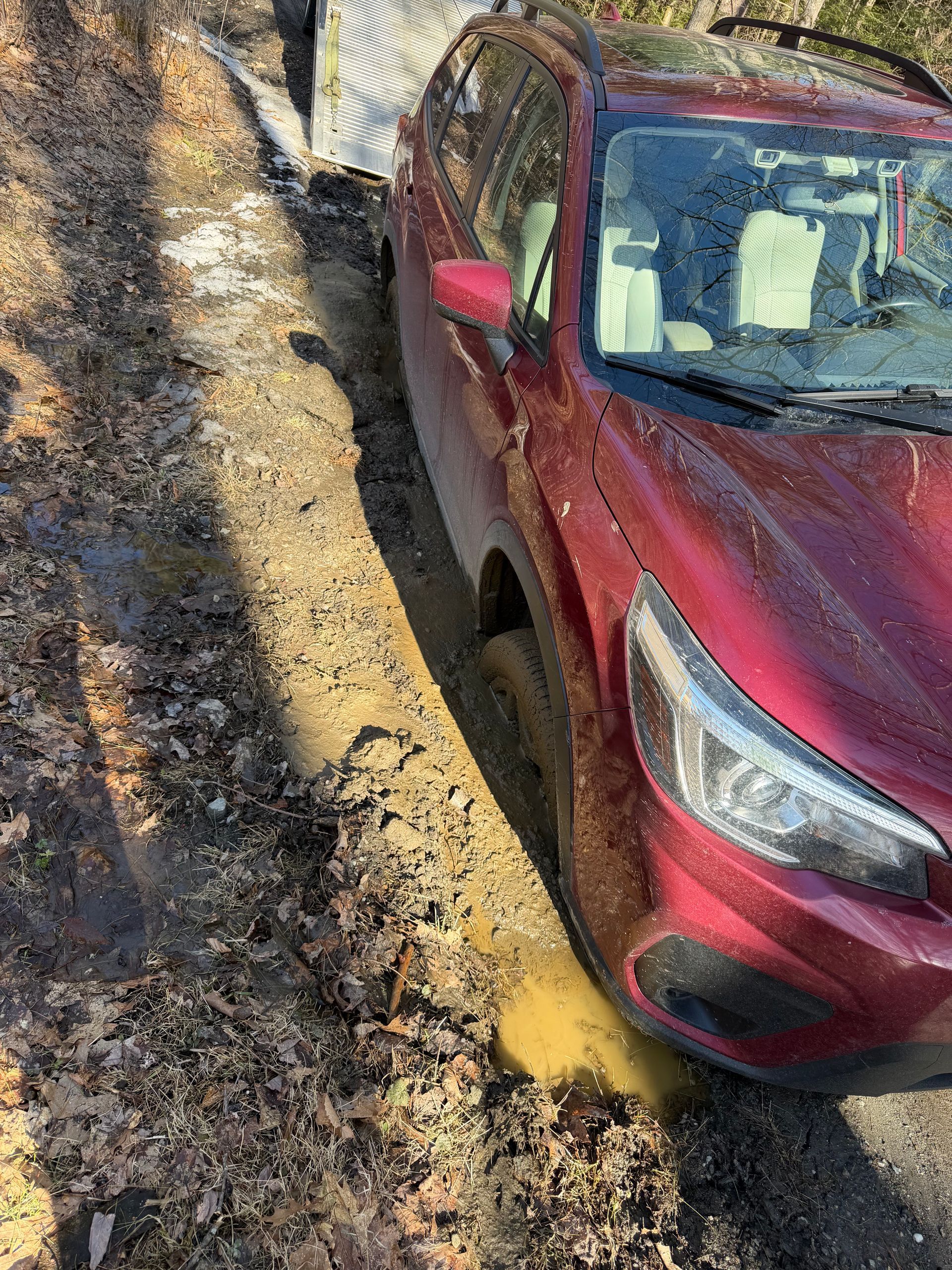 A red SUV is stuck in deep, muddy ruts along the side of a dirt path near a metal fence. MAACS Tow LLC