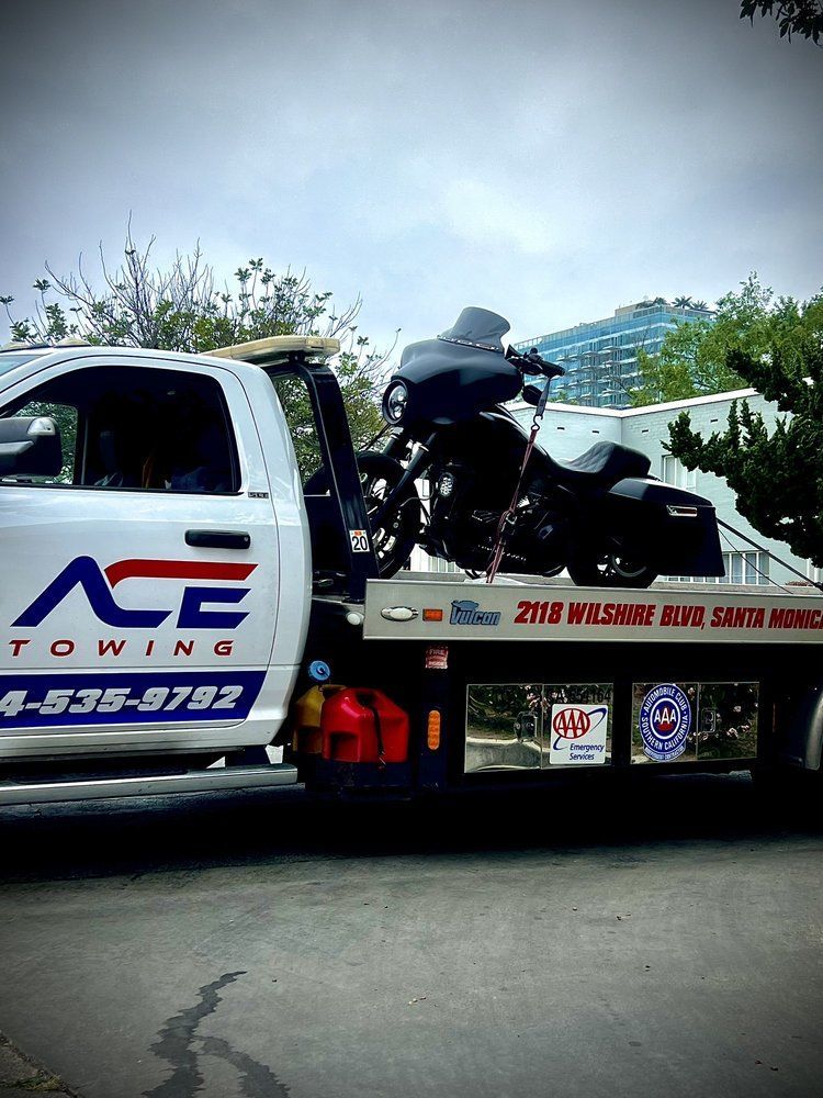 Tow truck carrying a black motorcycle, with “ACE TOW” logo on the cab and a flatbed trailer.
