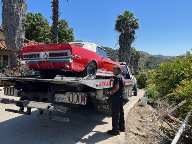 Red classic car on a tow truck beside a road, with a person standing nearby and palm trees in the background
