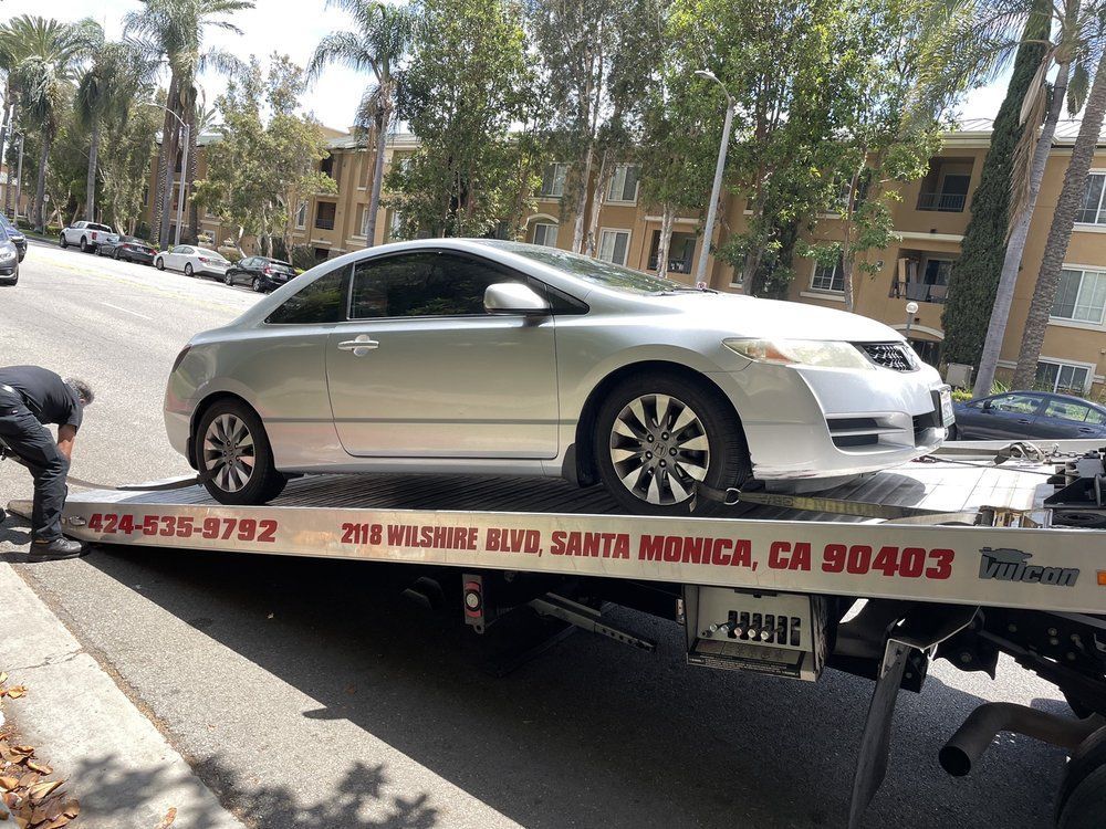 Silver sedan on a tow truck on a street lined with trees and apartment buildings.