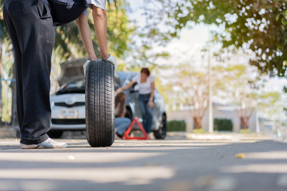 Person changing a car tire on a suburban street, with a car and another person in the background.