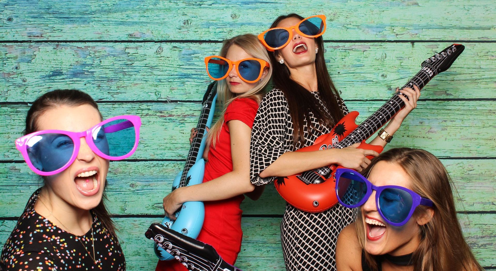 Vier Personen mit riesigen Sonnenbrillen und aufblasbaren Gitarren posieren vor einer blauen Holzkulisse.