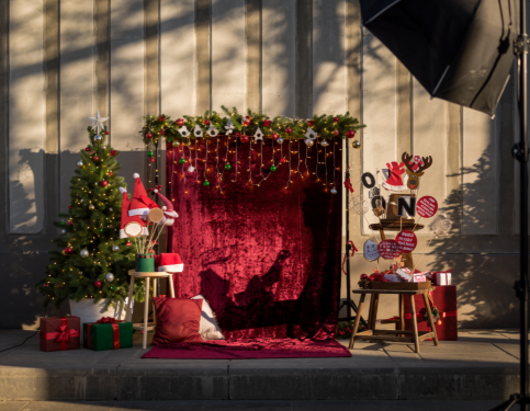 Christmas photo backdrop: tree, red velvet drape, garland, Santa hat, gifts, and a light setup.