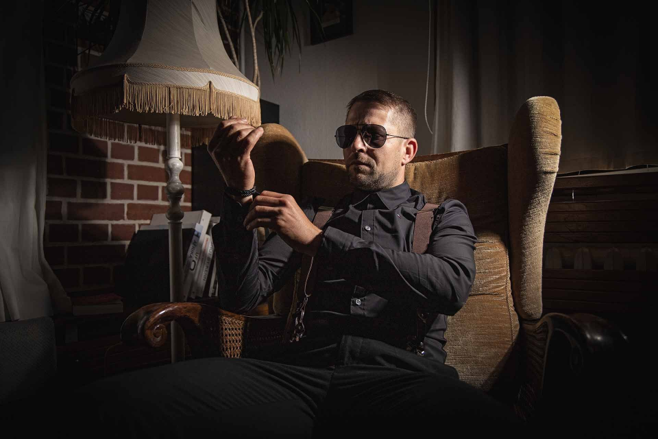 Ein Mann mit Sonnenbrille sitzt in einem Vintage-Sessel und hält einen Hut. Dunkle Umgebung, Lampe im Hintergrund.