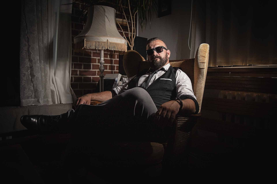 Ein Mann in Weste und Sonnenbrille lehnt sich in einem schwach beleuchteten Raum im Schein einer Lampe in einem Sessel zurück.