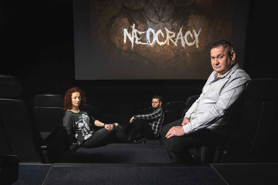 Drei Personen sitzen in einem dunklen Theater; auf der Leinwand erscheint der Titel „Neokratie“.