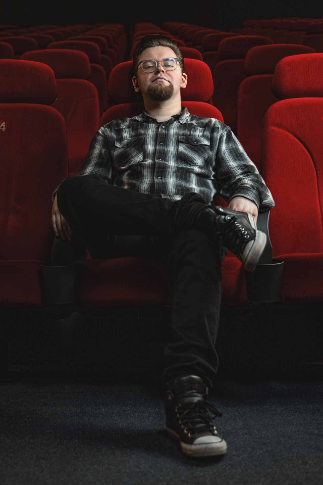 Ein Mann sitzt entspannt in einem roten Kinosessel; er trägt ein kariertes Hemd, Jeans und Turnschuhe.