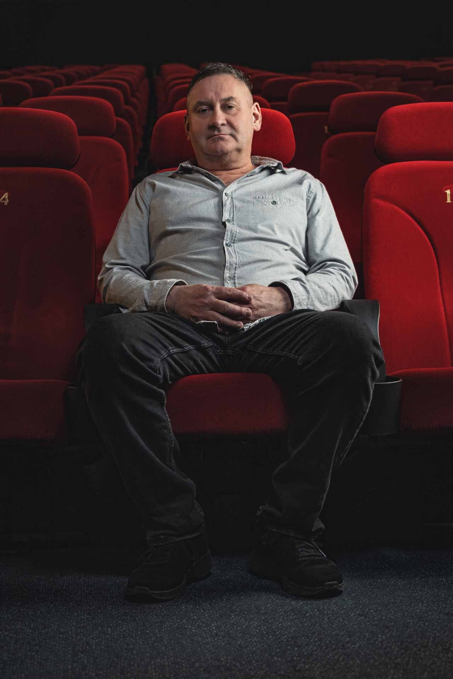 Ein Mann sitzt mit verschränkten Händen in einem roten Theatersessel. Er trägt ein graues Hemd und eine dunkle Hose.