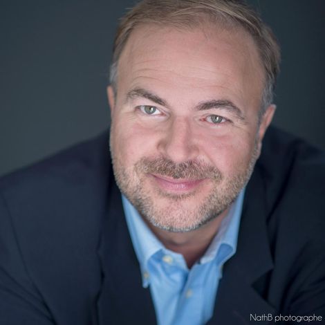 Portrait en gros plan d'un homme souriant, vêtu d'un blazer bleu marine et d'une chemise bleu clair, sur fond sombre.