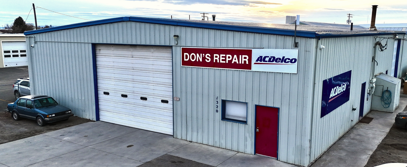 Exterior of Don's Repair auto shop; blue-gray metal building, red door, sign with red text, large garage door, blue car | Don's Repair