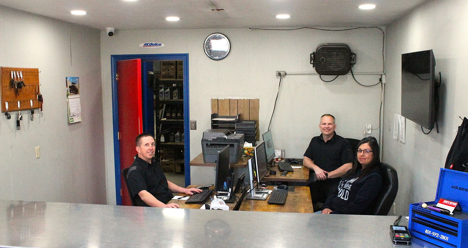 Three people at a desk in an auto shop office; one seated at a computer, two standing, with tools visible in the background | Don's Repair