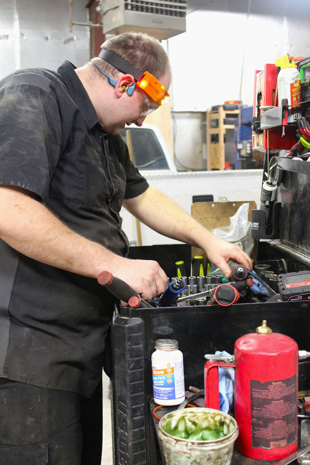 Mechanic wearing safety glasses, reaching into a toolbox. Tools, bottles, and a propane tank are visible in the shop | Don's Repair