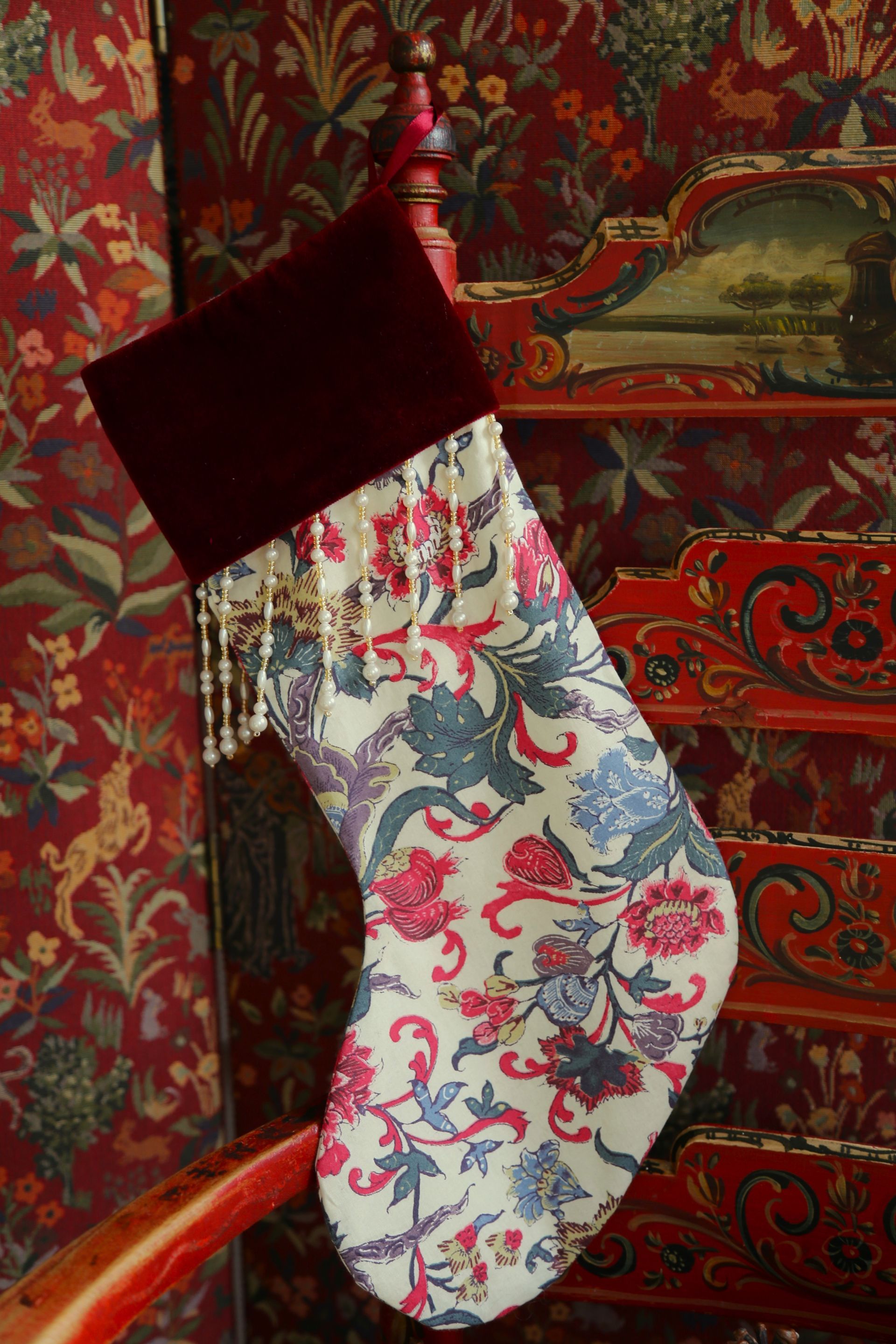 Een kerstsok met bloemenprint, een bordeauxrode fluwelen boord en kralenfranjes, hangend aan een rood geverfde trapleuning.