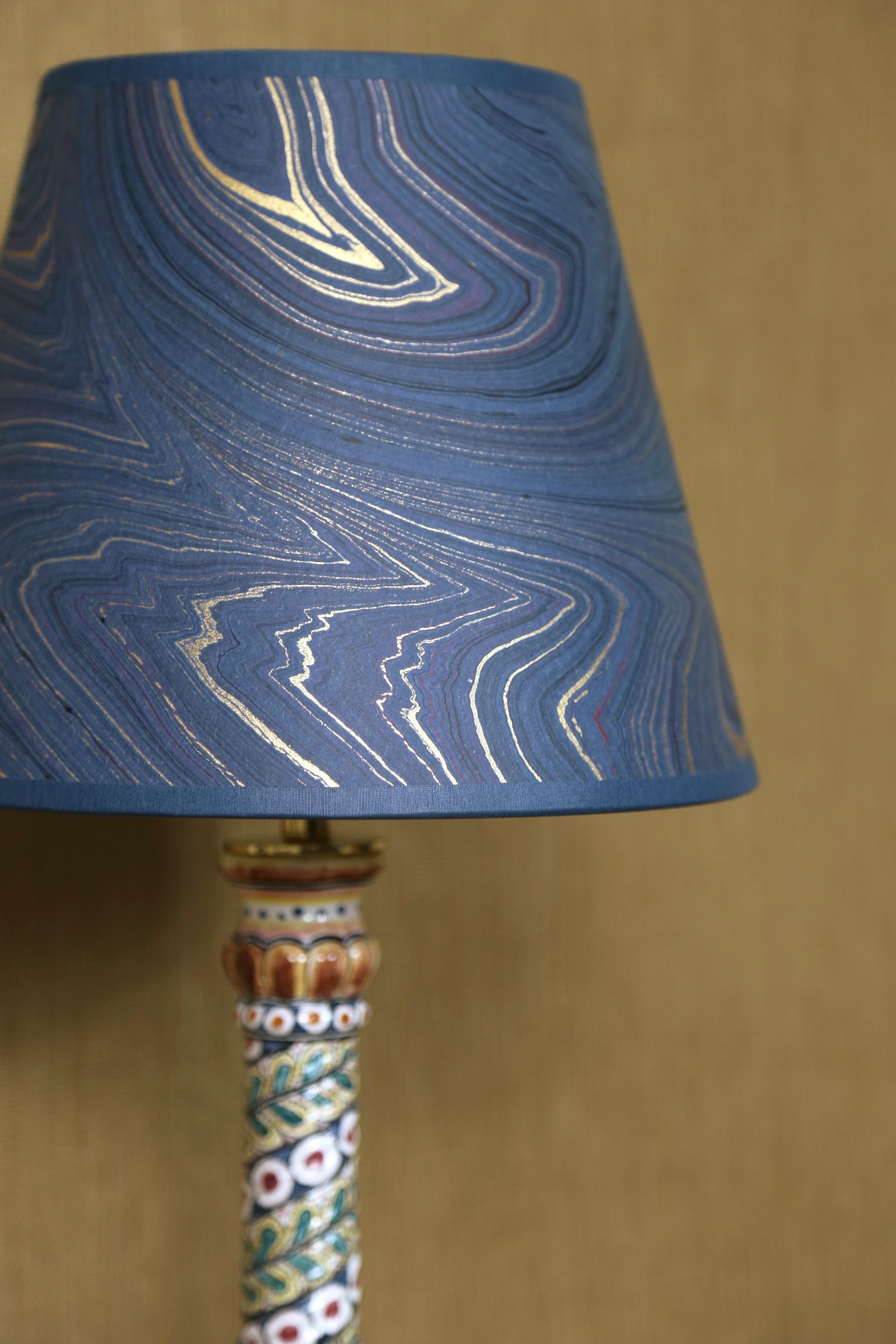Blauwe lampenkap met gouden marmering, geplaatst op een lampvoet met bloemenmotief, tegen een neutrale muur.