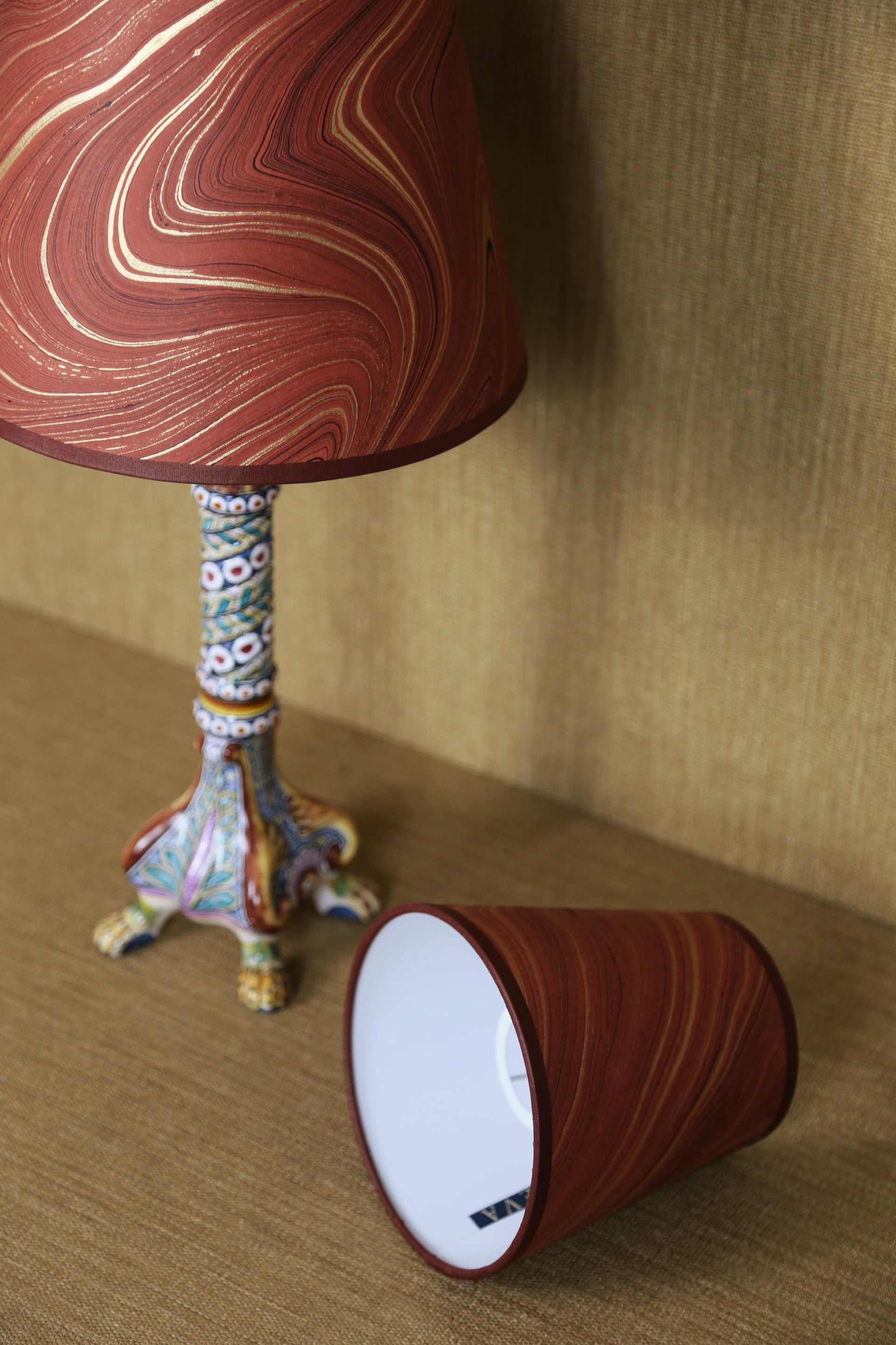Een tafellamp met een rood en goudkleurig patroon op de lampenkap, met een bijpassende, omgevallen lampenkap op een tafel.