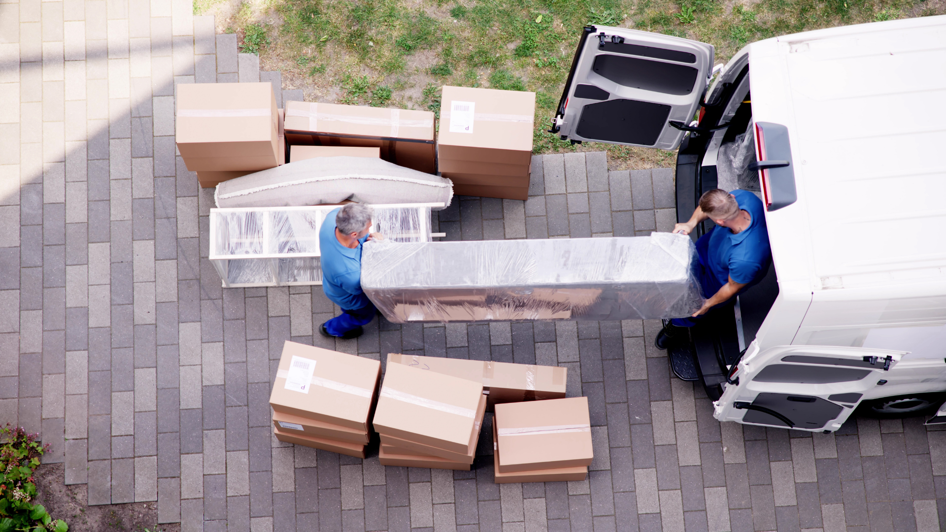 Two people in blue unload furniture from a van onto a brick patio, with several cardboard boxes nearby.
