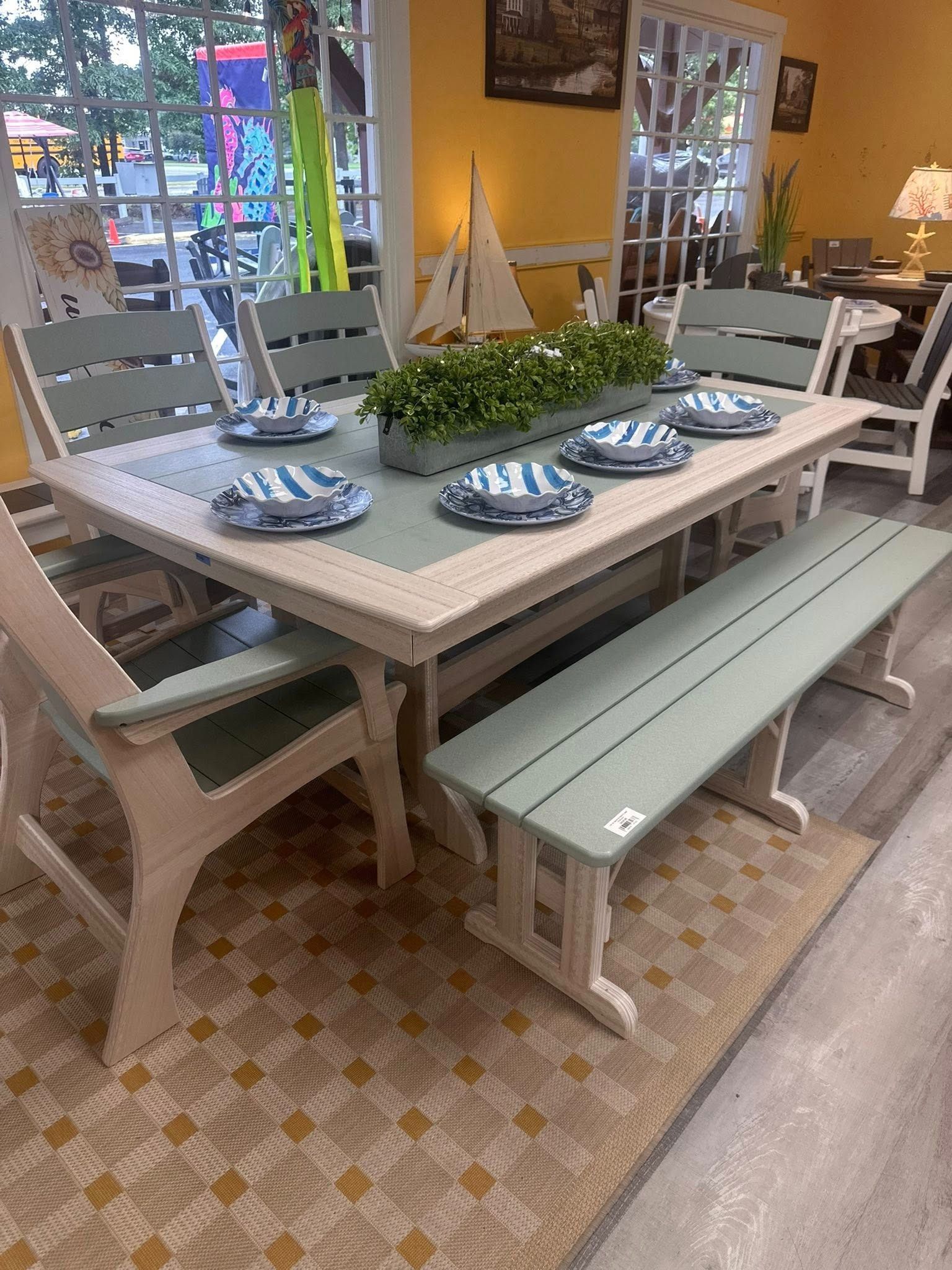 Outdoor dining set: cream and green table, chairs, bench, set for six with blue plates, flower box, on a rug.