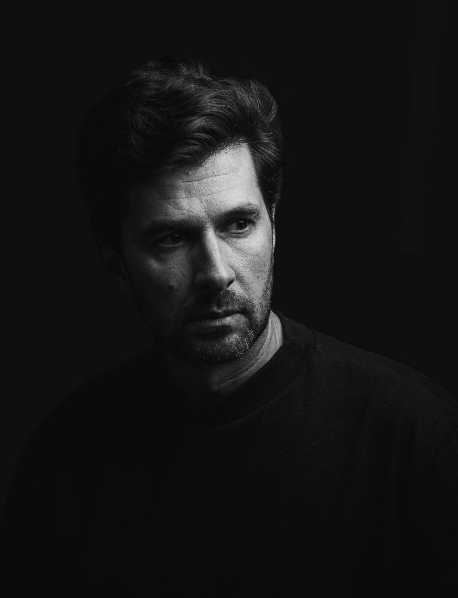 Zwart-wit portret van een man in een donker shirt, van opzij belicht tegen een donkere achtergrond.