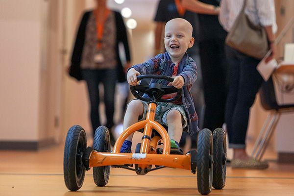 Een peuter lacht op een oranje driewieler binnenshuis, met wazige mensen die op de achtergrond lopen.