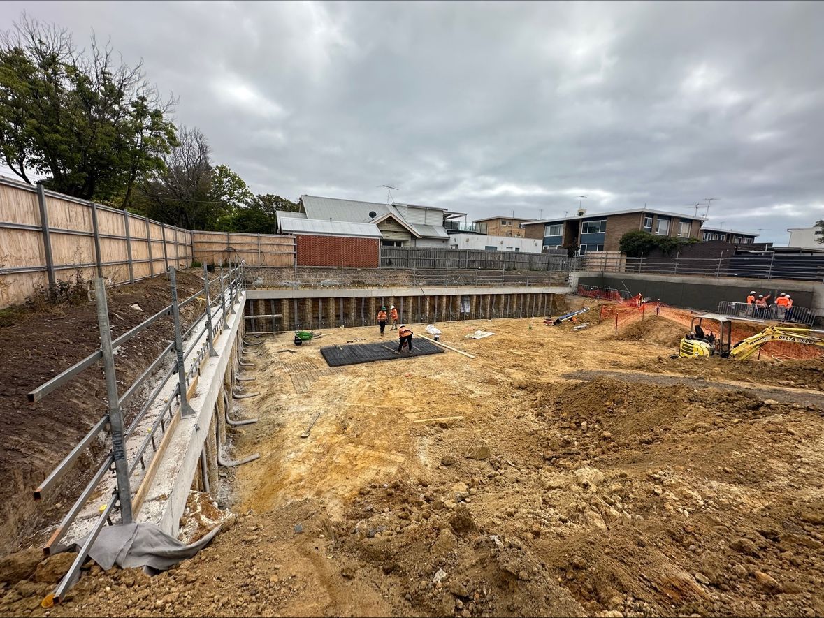 Construction site: excavated earth, retaining walls, workers, temporary fencing, overcast sky.