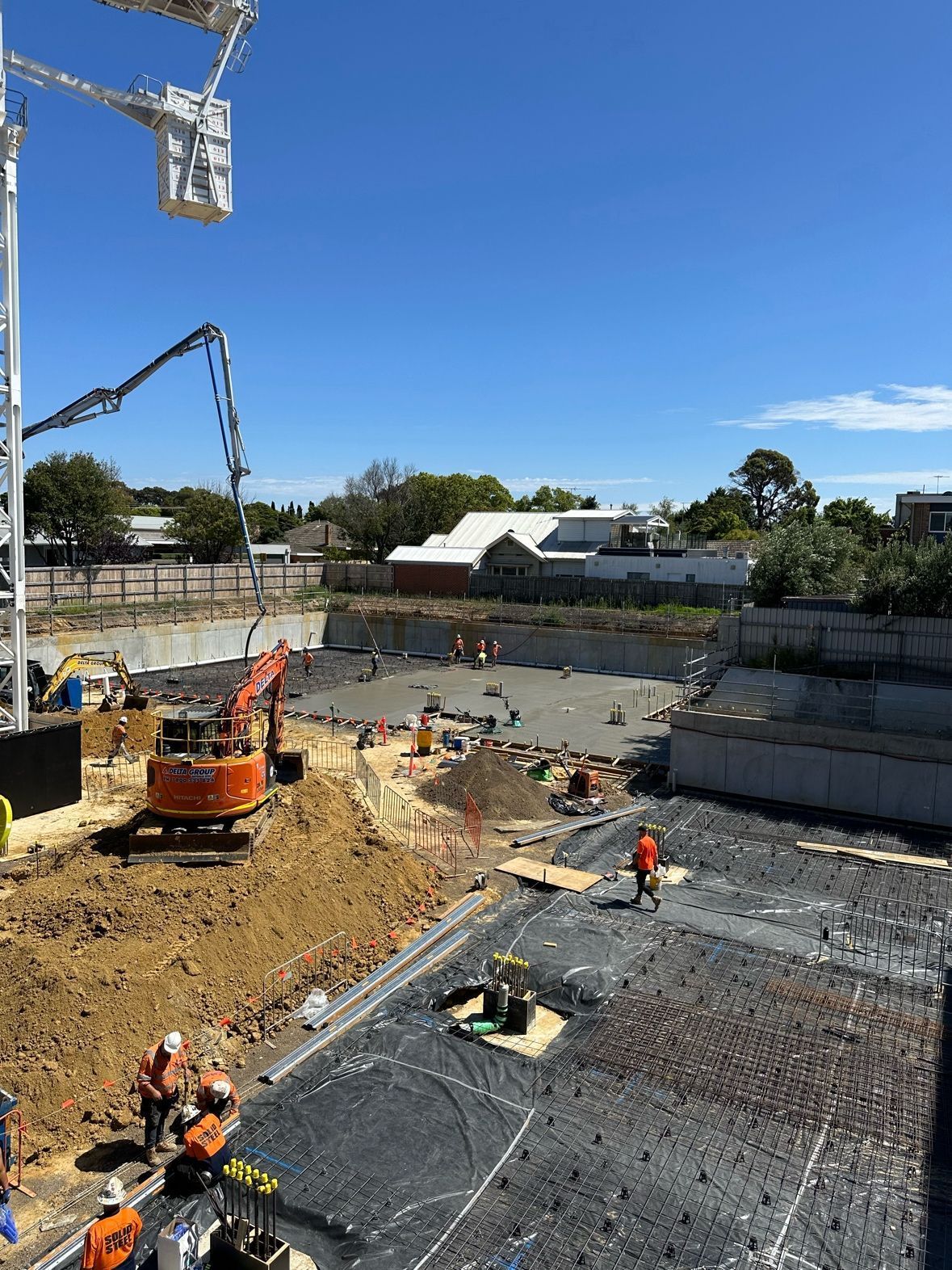 Construction site with crane, excavators, and workers. Orange safety vests, blue sky, concrete foundation.