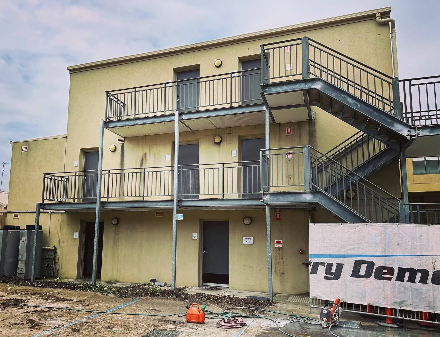 Two-story building with exterior metal stairs and balconies. Yellow walls, grey metal, and gray sky. Construction site.