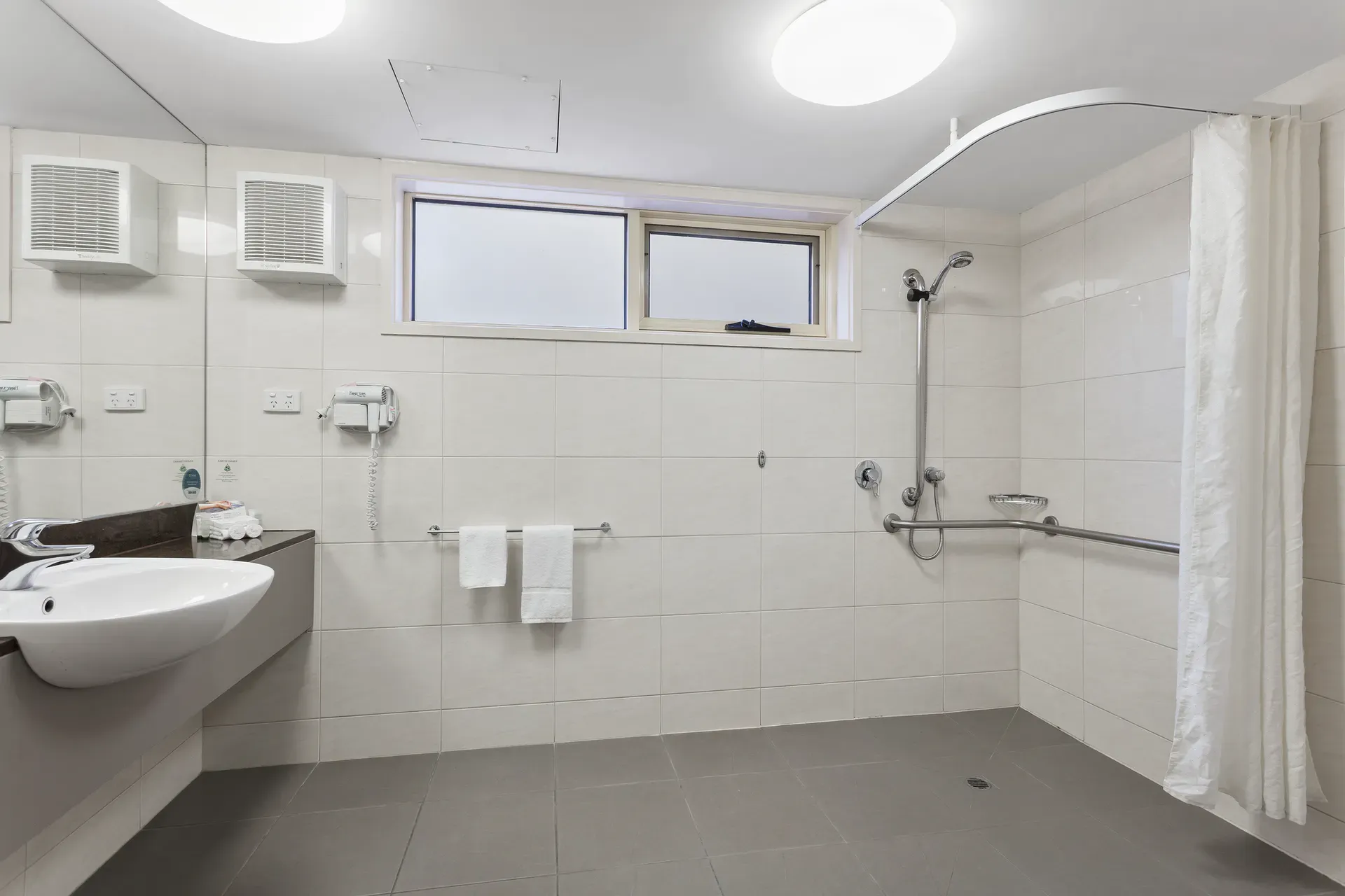 Accessible bathroom with shower, sink, and safety bars. White tiles and gray floor.