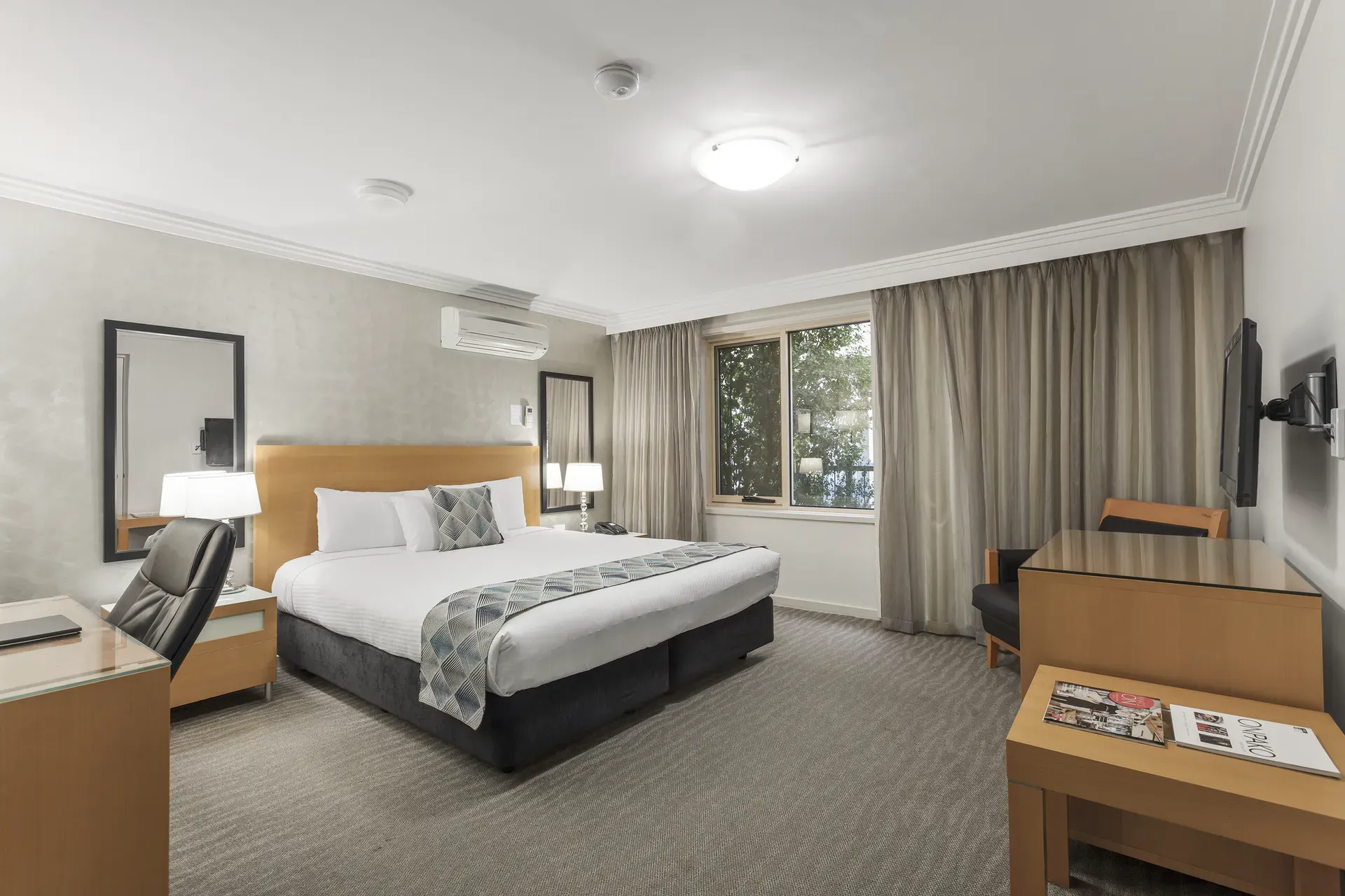 Hotel bedroom with a king-size bed, desk, and large window with curtains. Neutral colors.